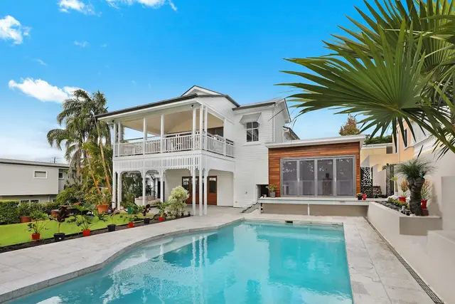 A backyard swimming pool beside a white two-story house and palm trees